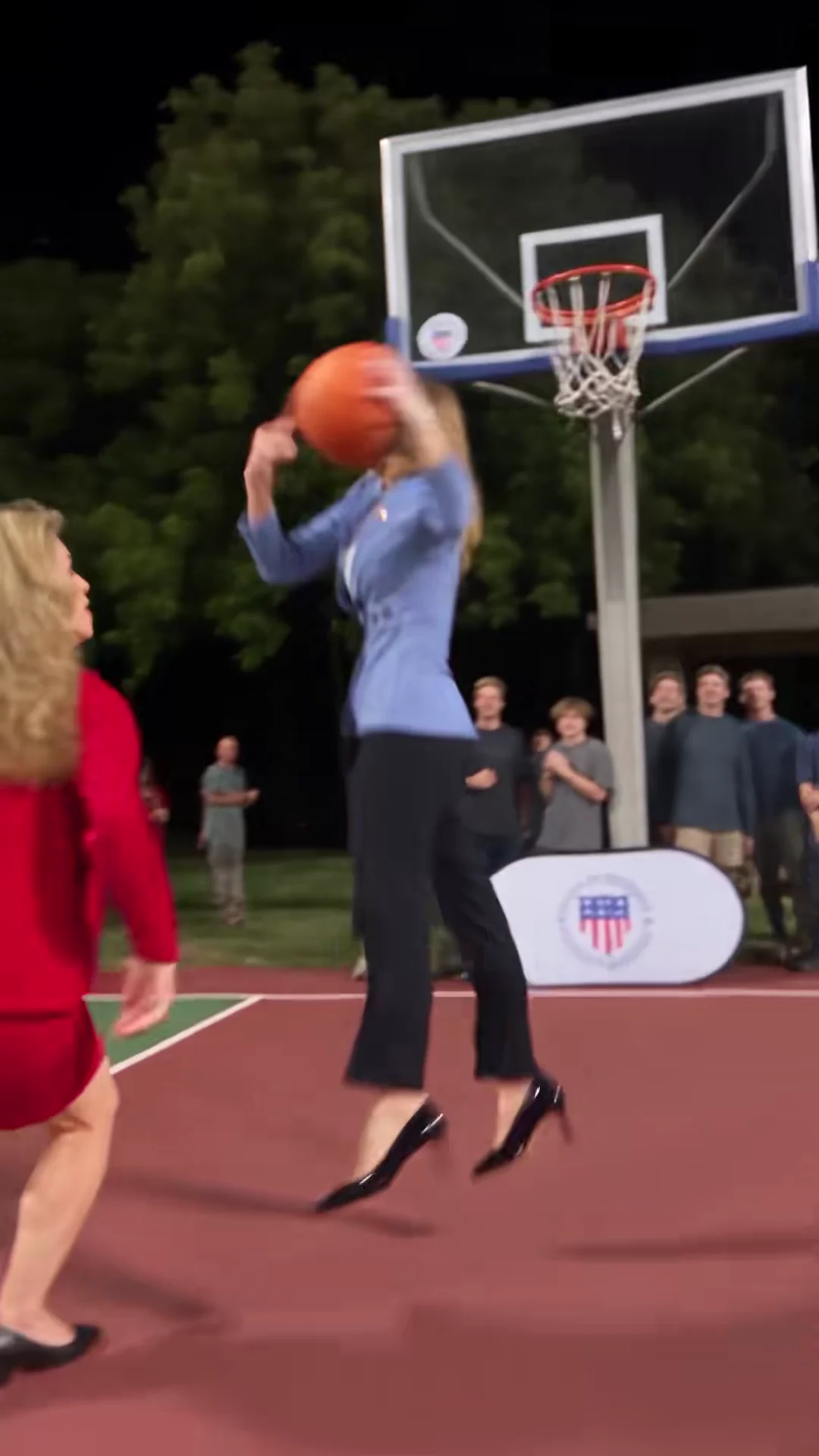 Credited source frame at about 7.2 seconds: the attacker in the light-blue blazer rises toward the rim with the ball over the defender in red, seen from behind, with spectators behind the hoop