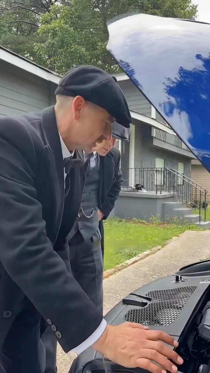Reference clip frame at about one second: a taller man in a dark flat cap, white shirt, black bow tie and long black overcoat bends over the open hood of a black car with a hand on the plastic engine cover, while a second man in a flat cap and grey three-piece suit stands partly hidden behind him, with a grey house and lawn beyond and the blue hood edge at the top right
