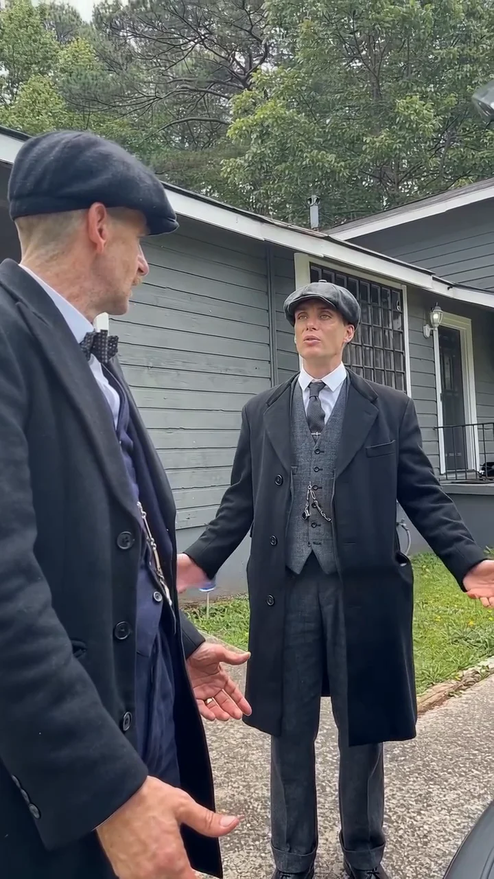 Reference clip frame at about twelve and a half seconds: a man in a flat cap, grey three-piece suit, tie and long black overcoat stands in the yard with both arms spread wide, while a taller man in a flat cap, bow tie and black overcoat listens in profile at the left edge with an open hand out
