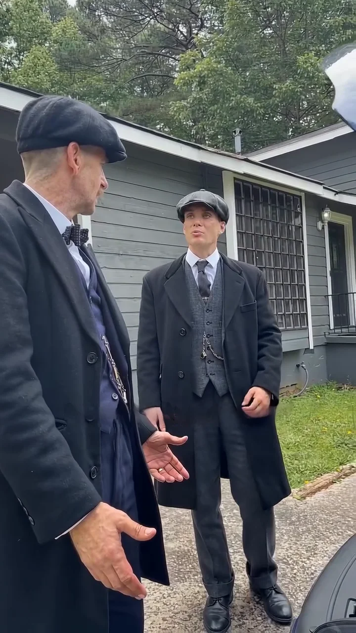 Reference clip frame at about eight and a half seconds: a taller man in a dark flat cap, bow tie and long black overcoat stands in profile at the left edge of the frame with an open hand out, facing a man in a flat cap and grey three-piece suit who stands in the middle of the frame in front of a grey house with a barred window, with his hands low at his sides