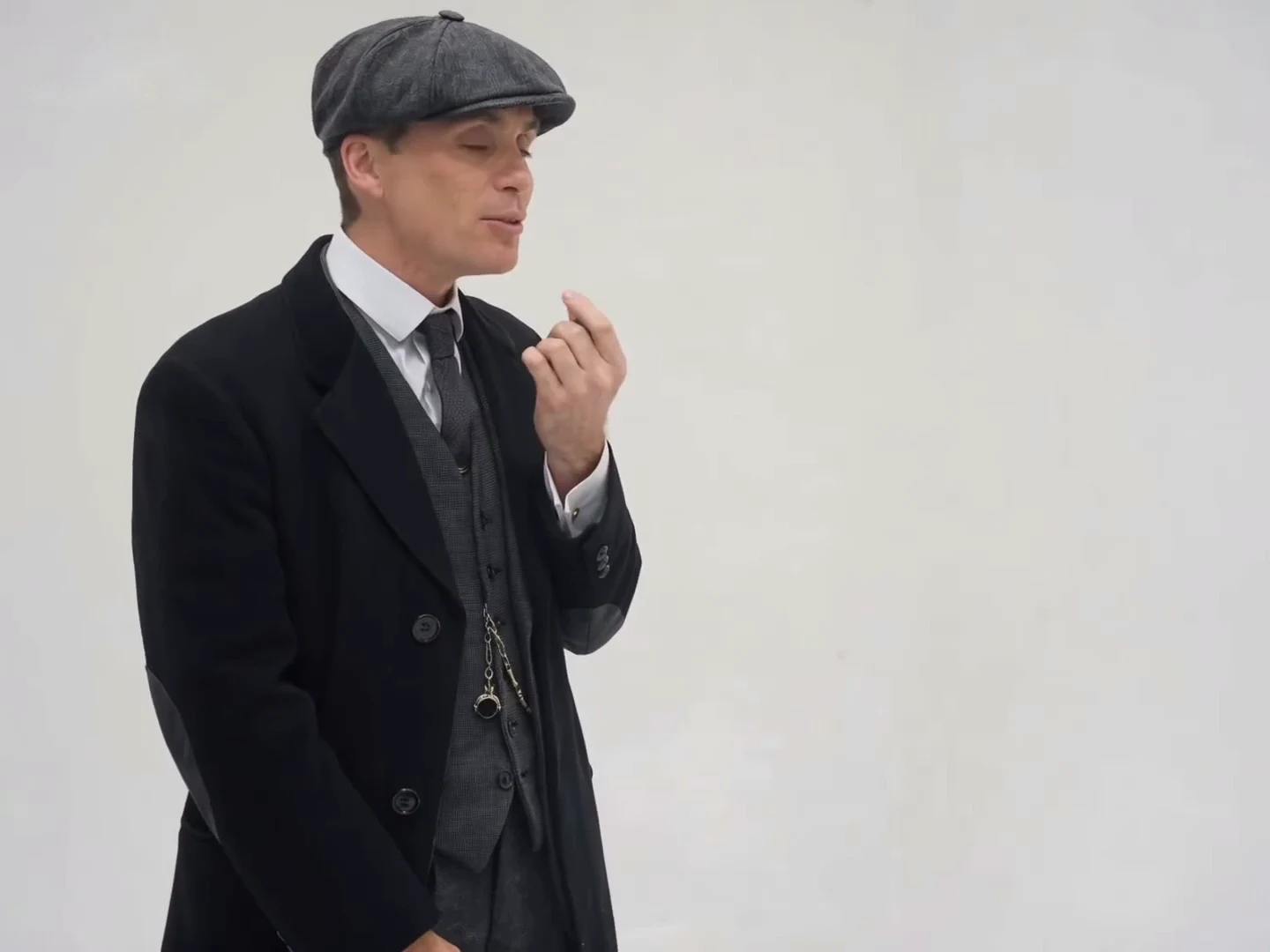 Credited source post frame at about thirteen seconds: the man in the flat cap and overcoat in the slightly closer framing, holding the small black device