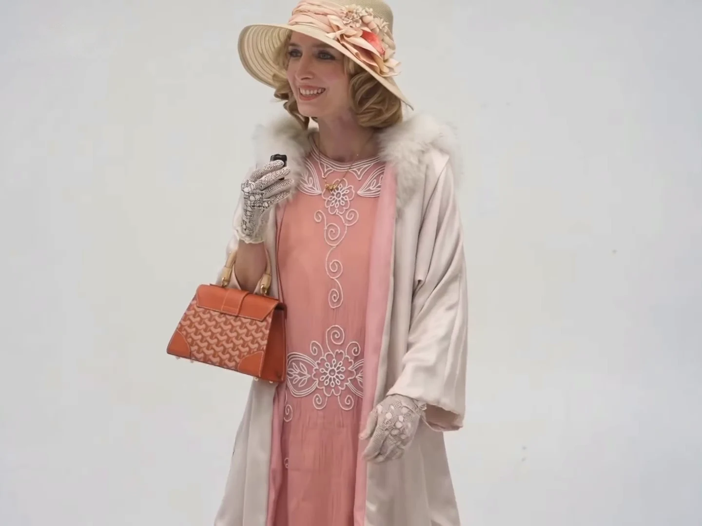 Credited source post frame at about five and a half seconds: a blonde woman in a flowered wide-brim hat, pink dress, fur-collared coat and white gloves holds an orange handbag and a small black device near her chin