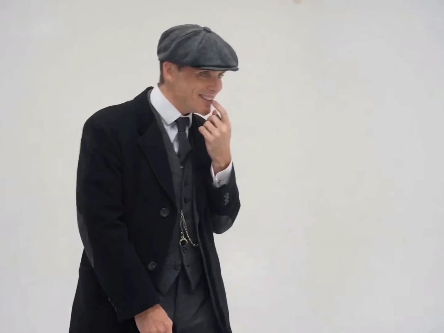 Credited source post frame at about seven and a half seconds: a man in a grey flat cap and black overcoat touches his lip with a small black device in his fingers, against a plain grey backdrop