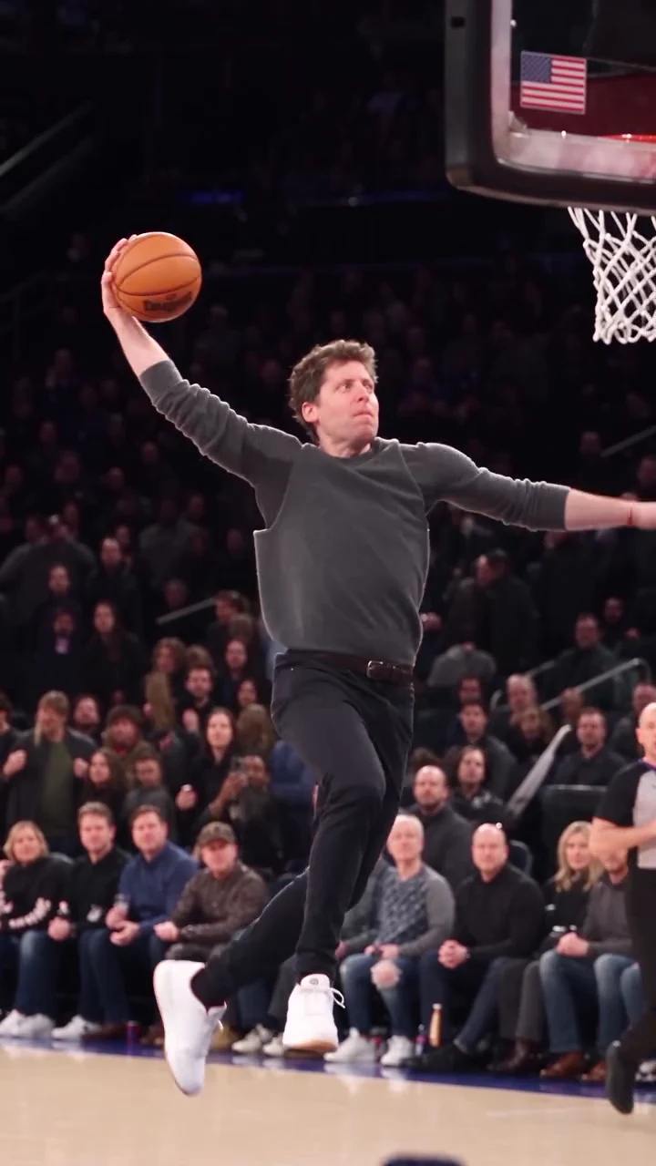 Reference frame at about ten and a half seconds: a slim man in a dark grey long-sleeve top and dark trousers is in mid-air under a glass backboard, one arm raised high with a basketball, the rim and white net at the top right and a packed crowd behind
