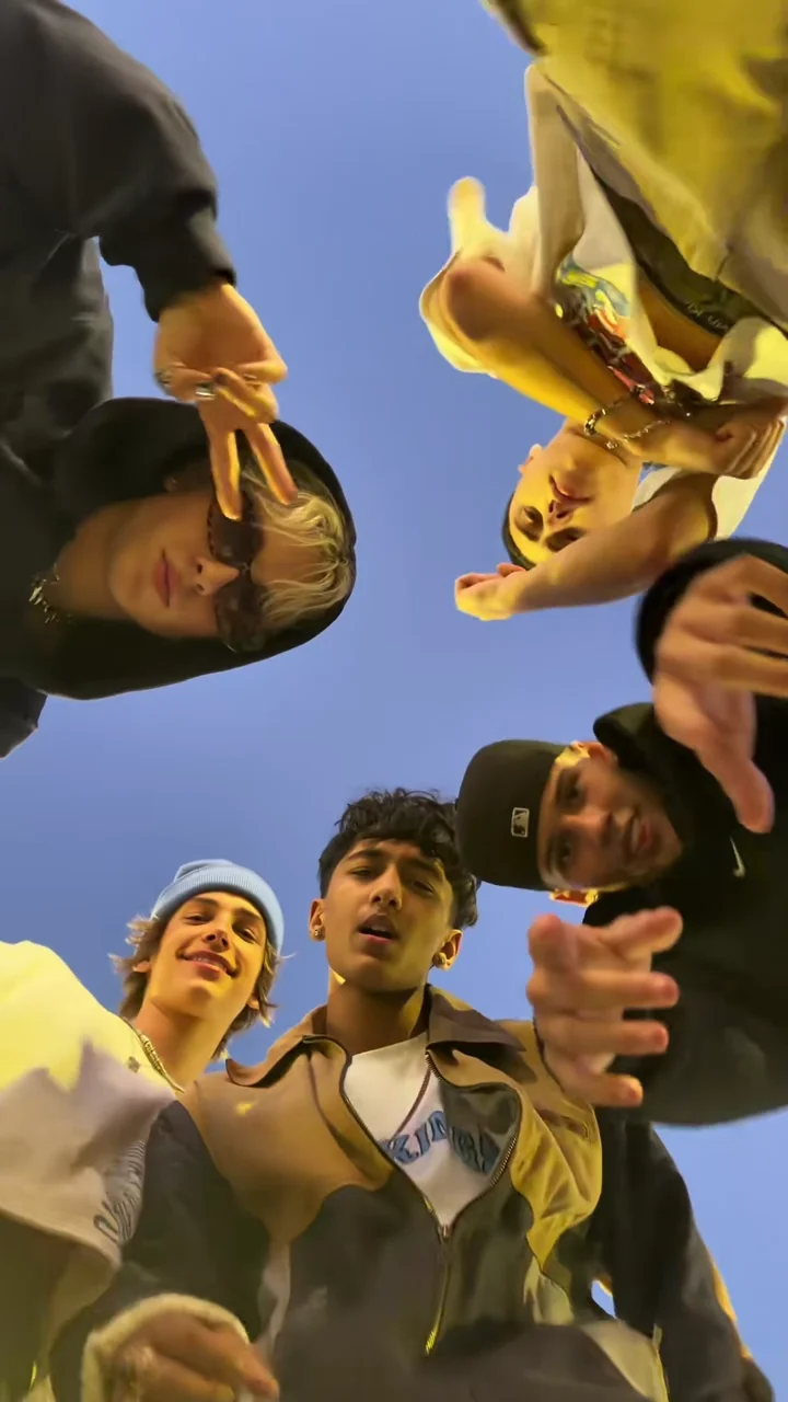 Credited source frame at about 0.4 seconds: five faces lean over a phone on the ground against a blue sky
