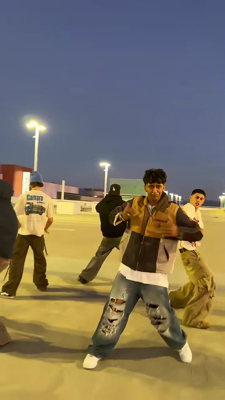 Credited source frame at about 5.5 seconds: the leader in a tan-and-navy patchwork jacket and ripped jeans dances front and centre on the rooftop car park with the others behind