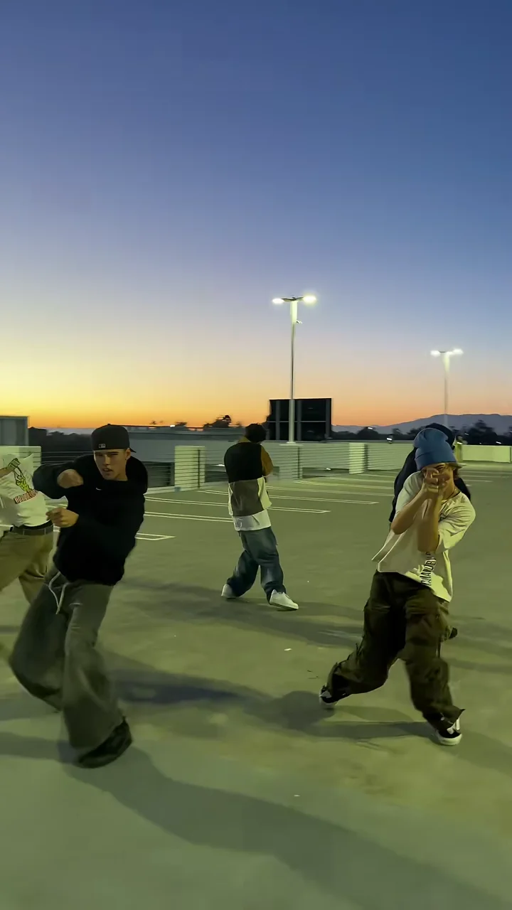 Credited source frame at about 10 seconds: the crew dances on a rooftop car park at sunset with light poles behind, a dancer in a black cap and one in a blue beanie in front