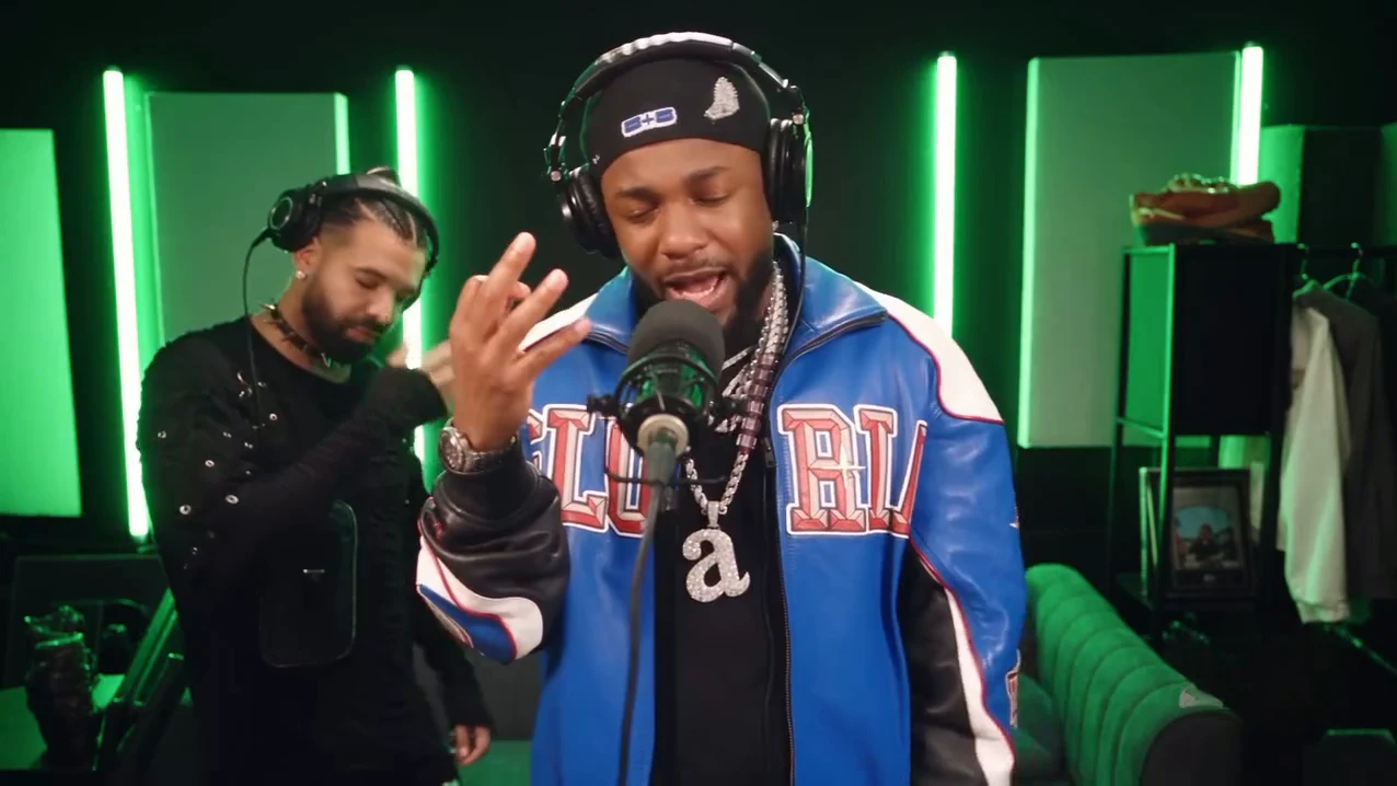 Reference frame at nine seconds: in a dark recording booth lit by vertical green light strips, a person in a black durag, studio headphones and a blue-and-white varsity jacket stands at a microphone with one hand raised, while a second person in black with braided hair and headphones stands behind on the left