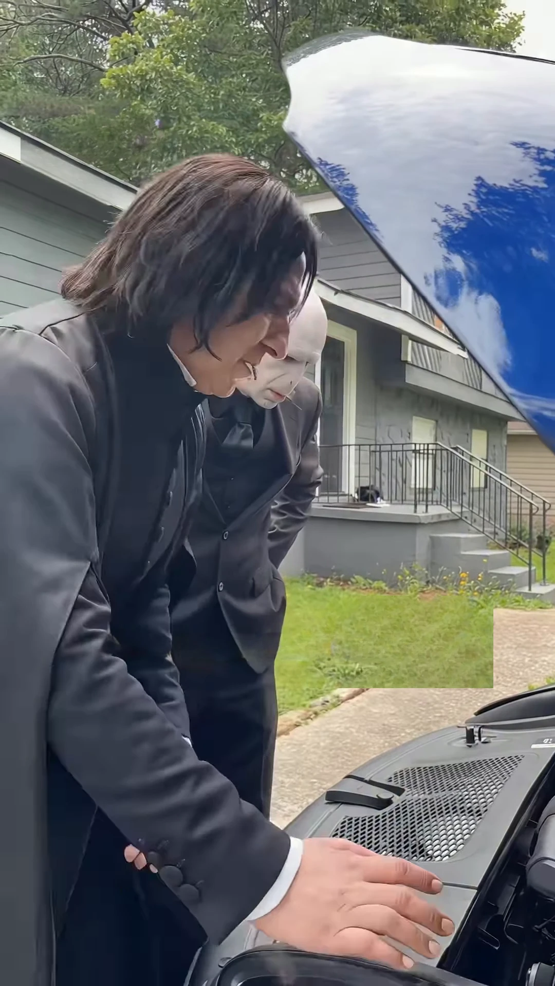 Credited source reel frame at about one second: a tall figure in a long black coat bends over the open hood of a black car and touches the plastic engine cover, while a bald figure in a black suit stands behind in a suburban yard