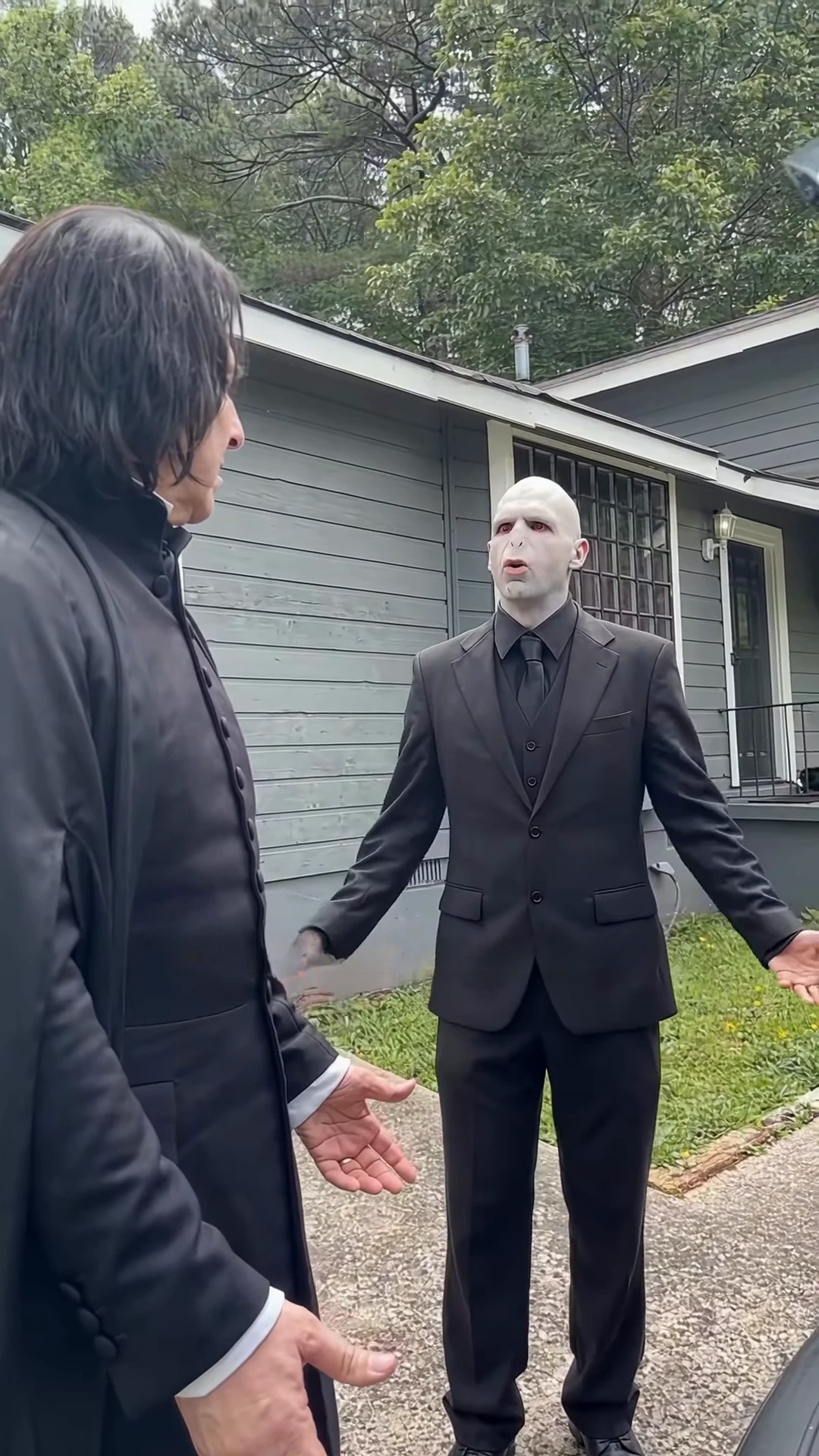 Credited source reel frame at about twelve and a half seconds: a bald figure in a black suit and tie stands in a yard with both arms spread wide while a long-haired figure in a black coat listens at the left edge