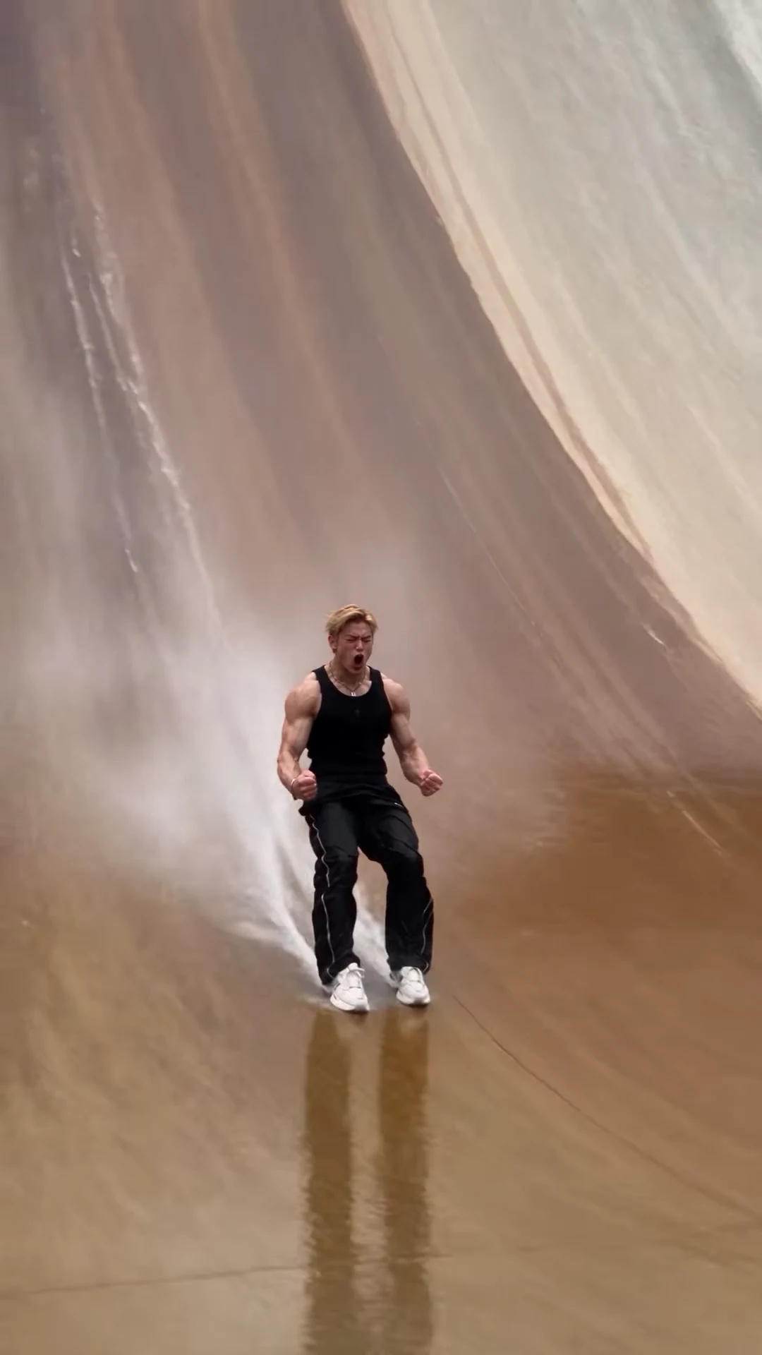 Reference frame at twelve seconds: a muscular man in a black tank top and black track pants slides down the wet, brown-streaked face of a curved concrete dam, fists clenched, with water spraying behind him