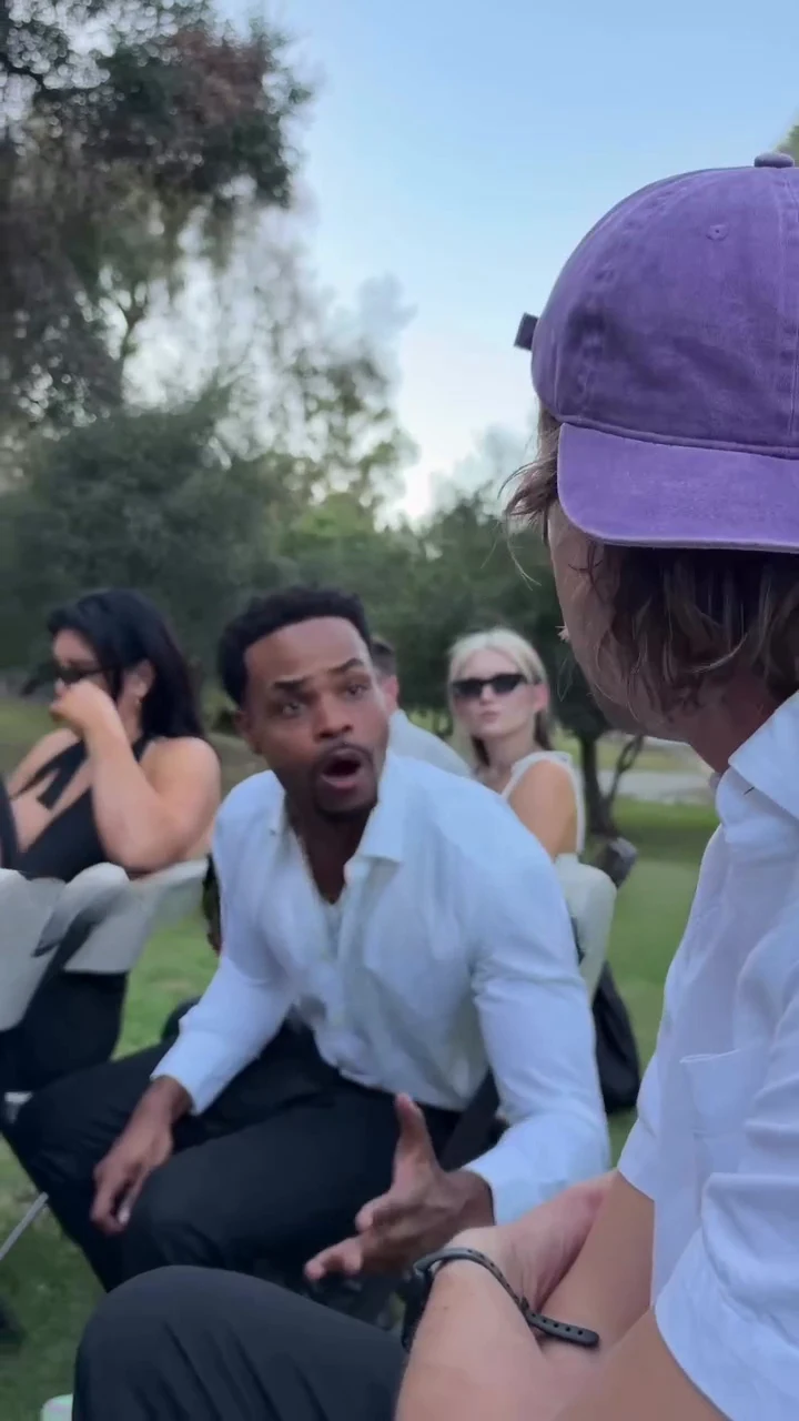 Reference frame at about five and a half seconds: on a lawn under oak trees, a man in a white open-collar shirt and black trousers sits on a folding chair leaning forward and gesturing with both hands, mourners in black with sunglasses behind him, and the back of a head in a purple cap in the right foreground