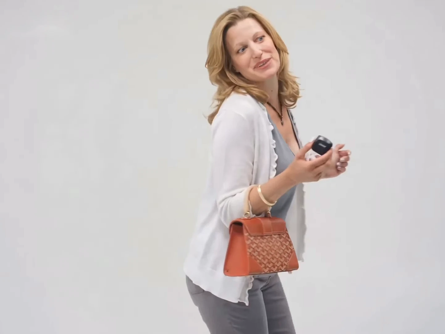 Credited source reel frame at about two and a half seconds: a blonde woman in a white cardigan over a grey top with an orange handbag looks over her shoulder, away from the small black device she holds, against a plain grey backdrop