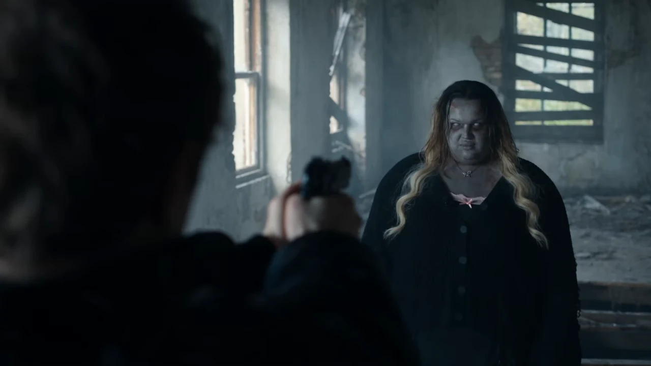 Reference frame from the room: seen over the survivor's shoulder, a pistol is raised in the foreground while a pale zombie with clouded white eyes, forehead cuts and long blonde-brown hair stands in a derelict grey room